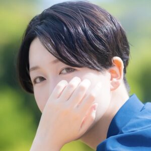 Close-up of a person with dark hair in a blue shirt, covering part of their face with a hand outdoors.